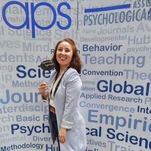 Photo of CUNY SPS Psych major Nurhan Michel posing at a background that reads "APS".